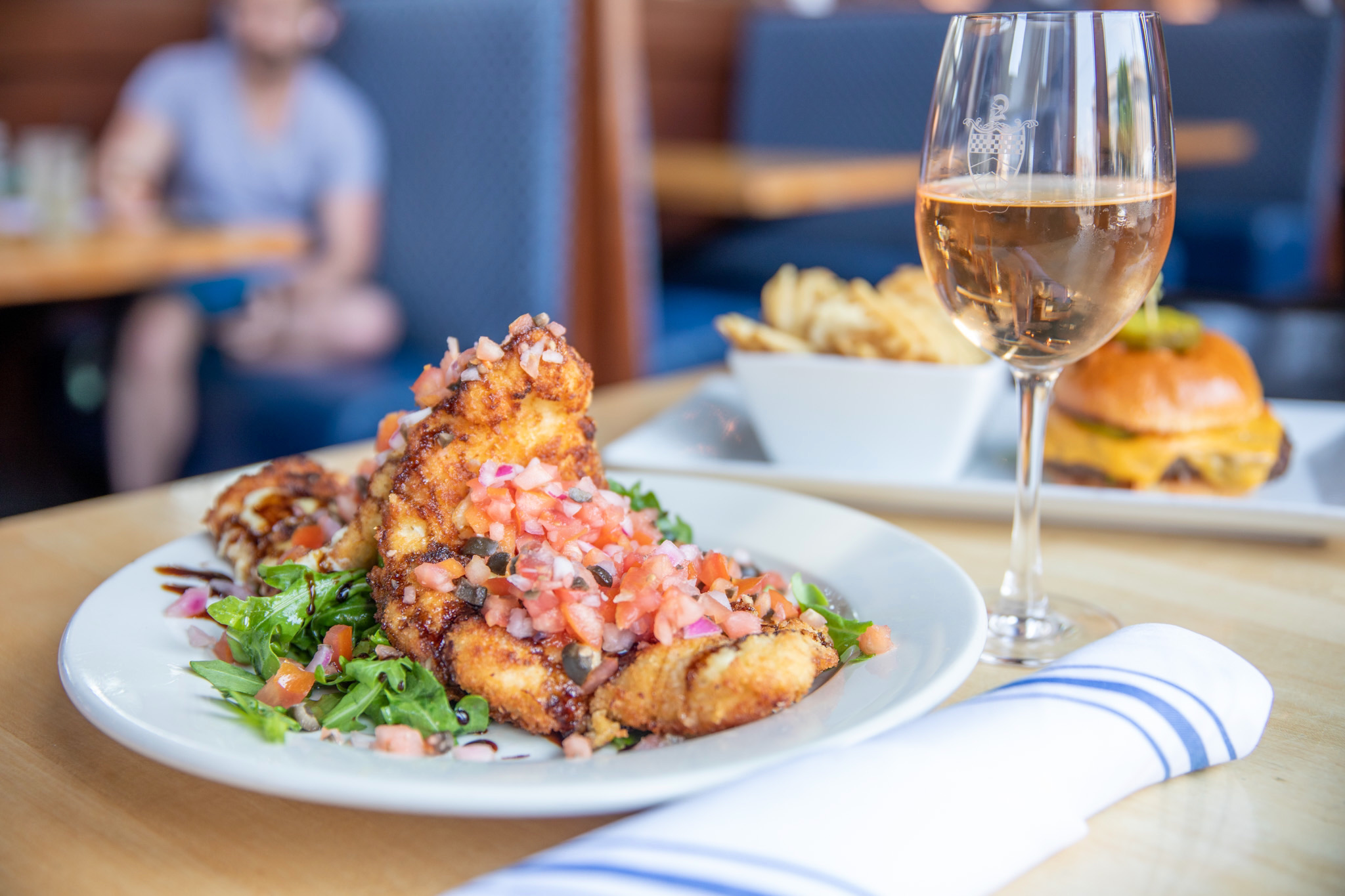 A plate of breaded chicken topped with diced tomatoes and herbs sits on a table alongside a side salad. A glass of wine complements the meal, while a menu displays fries and a burger in the background, all within the restaurant's casual atmosphere.