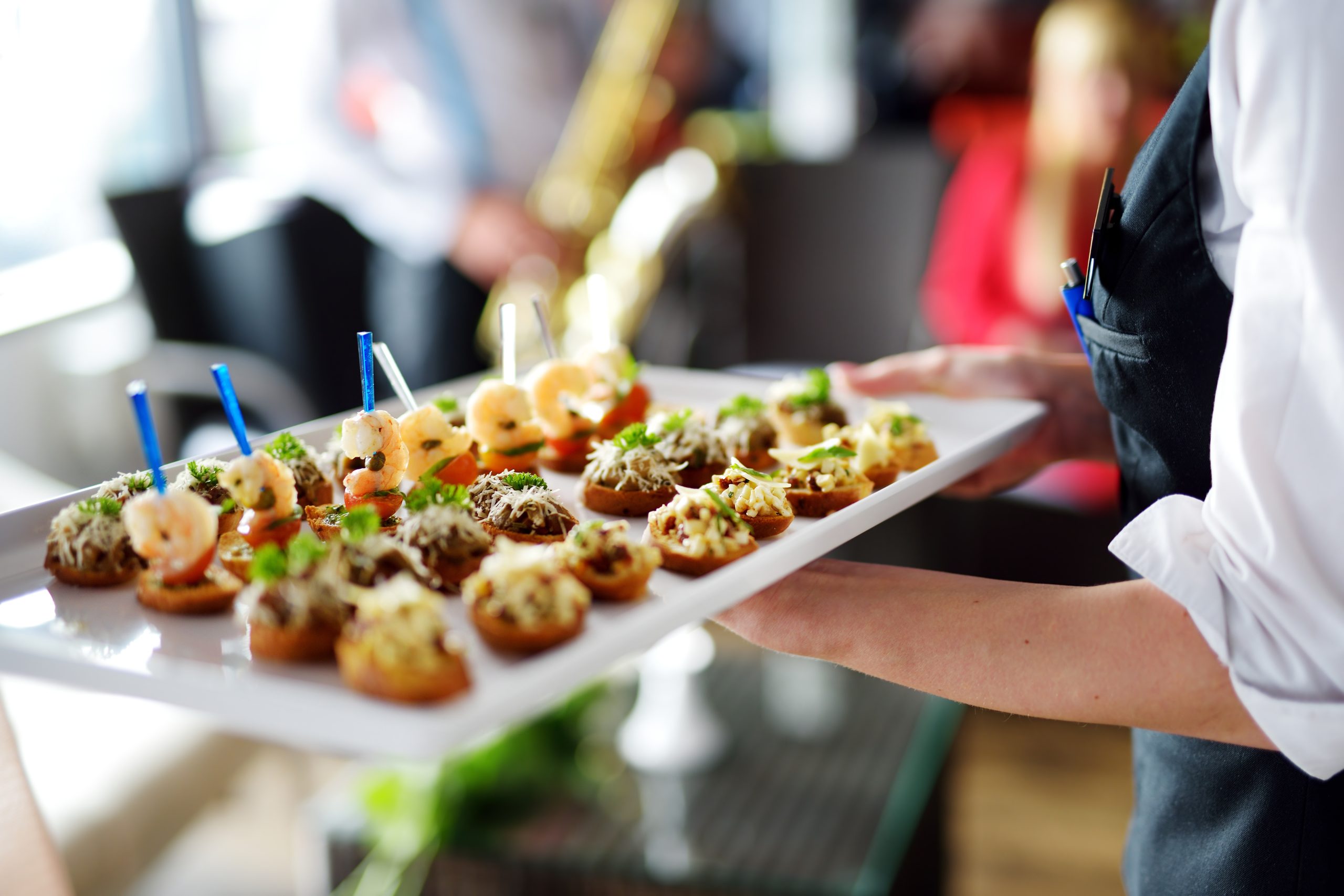 At a lively event, a server carries a tray of assorted appetizers, featuring shrimp and various toppings on small bread slices. In the blurred background, a musician plays a saxophone while guests mingle.