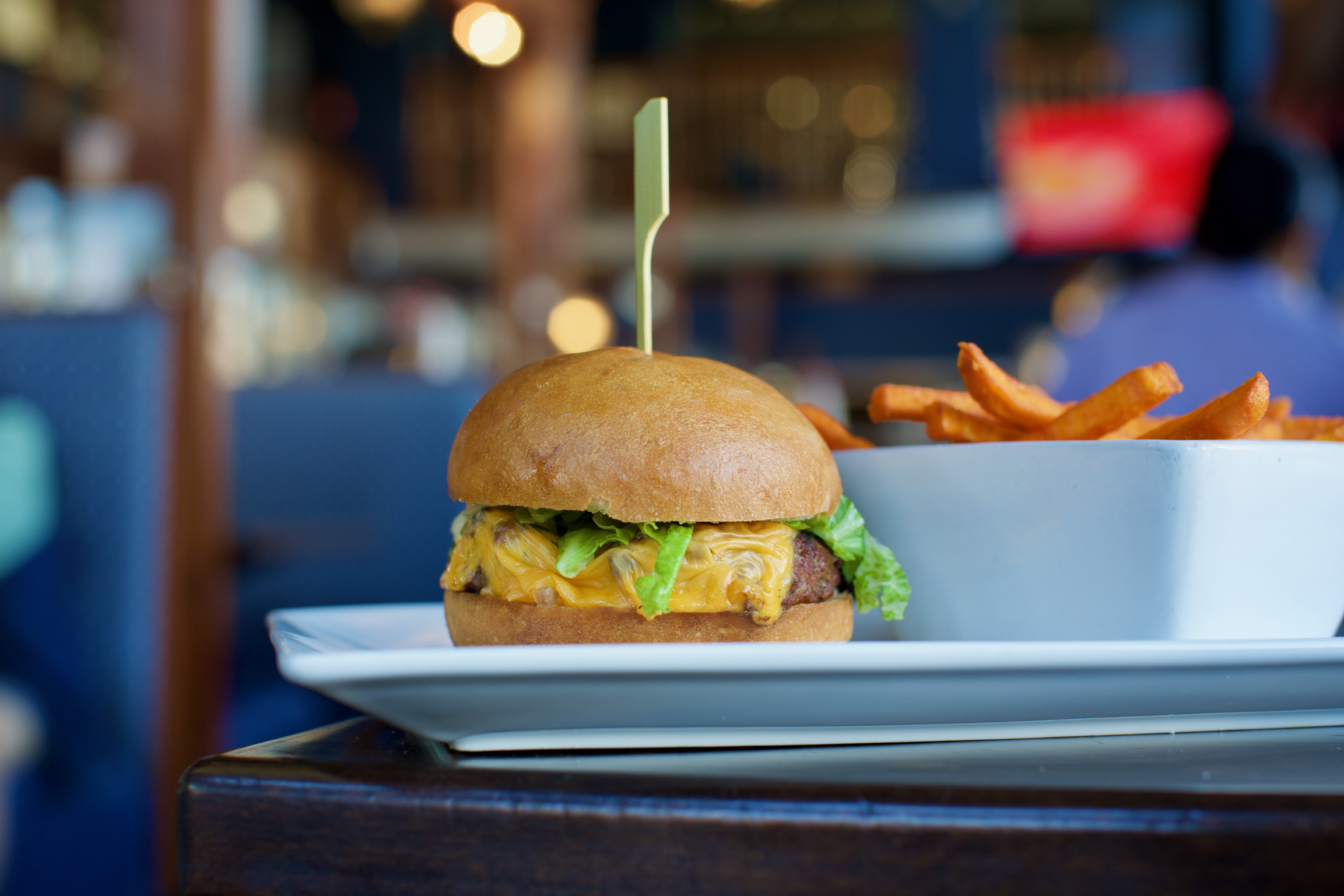 A cheeseburger with lettuce sits on a plate beside a bowl of sweet potato fries, inviting diners to explore the restaurant's carefully crafted menus. The blurred background, bathed in warm lighting, adds to the dish's enticing allure.