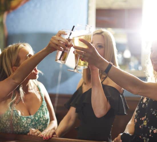 A group of women at a bar raise their glasses in a celebratory toast, marking the end of a delightful luncheon. Sunlight shines brightly through a window, creating a warm and joyful atmosphere. In the background, a mirror reflects the scene against the soft blue walls.