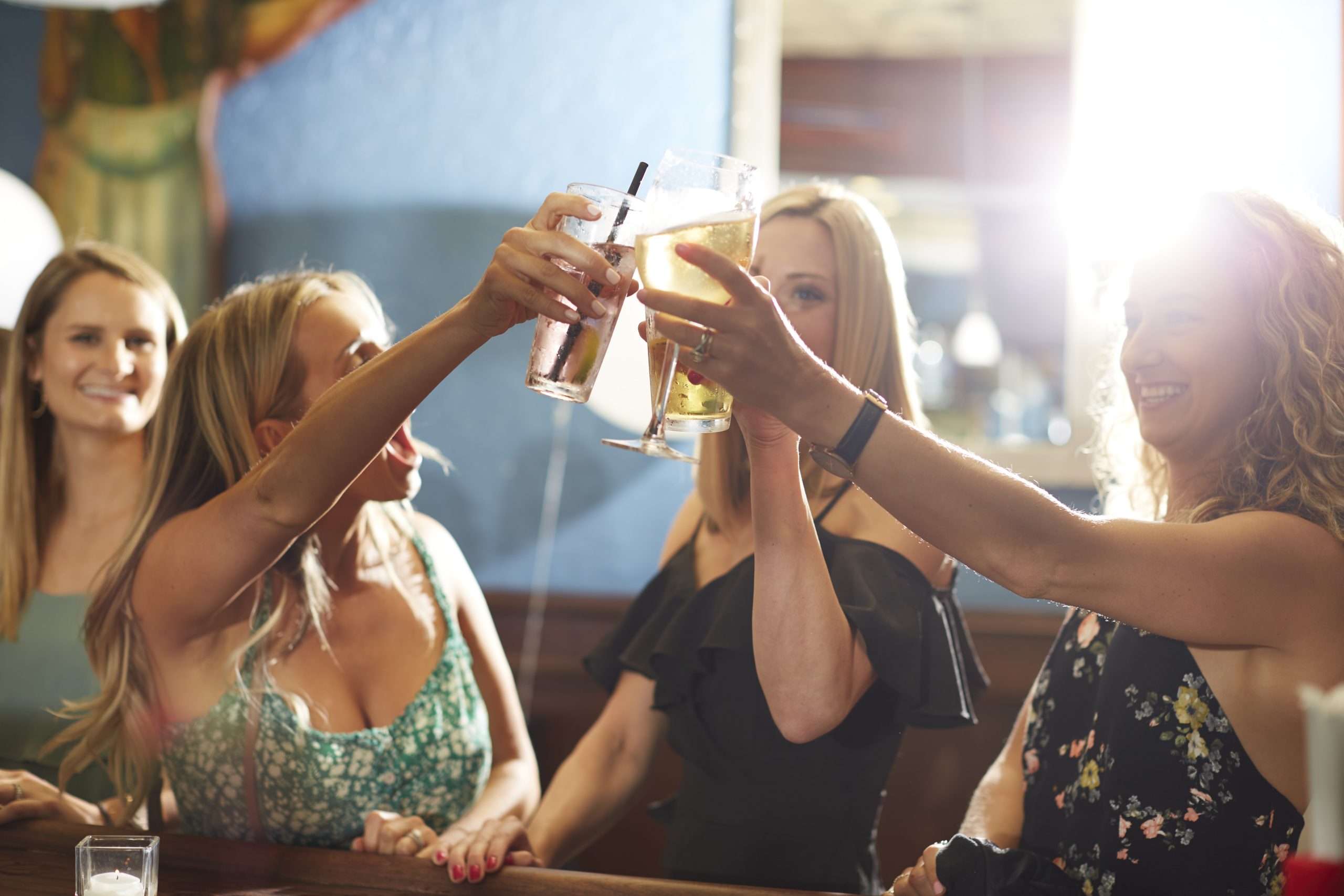 A group of women at a bar raise their glasses in a celebratory toast, marking the end of a delightful luncheon. Sunlight shines brightly through a window, creating a warm and joyful atmosphere. In the background, a mirror reflects the scene against the soft blue walls.