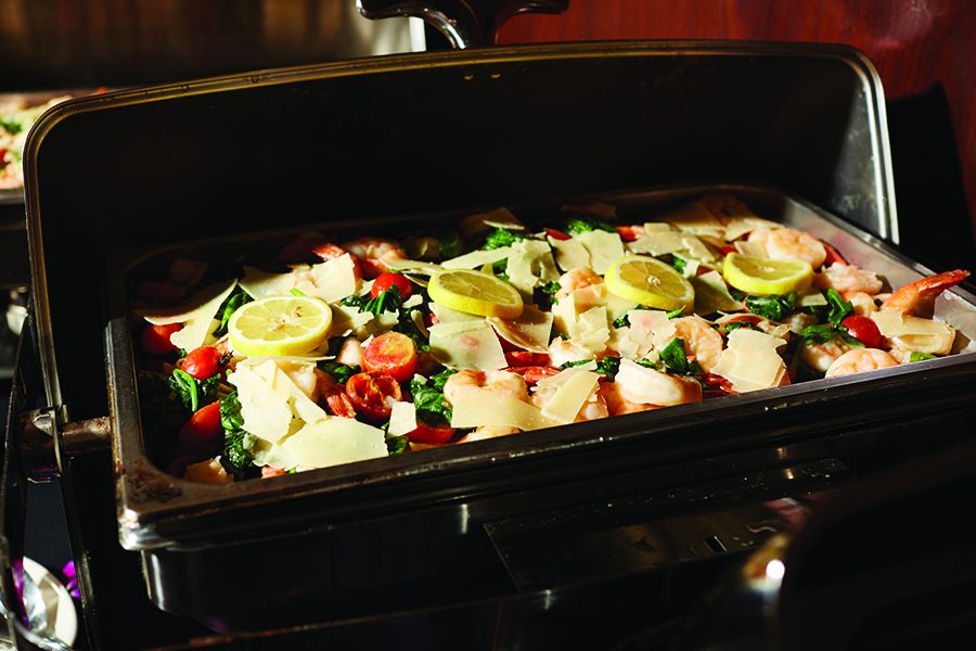 A buffet tray brimming with a colorful dish of shrimp, spinach, cherry tomatoes, and lemon slices takes center stage at events. Delicately garnished with cheese shavings and placed under a warm light, it offers a feast for both the eyes and the palate.