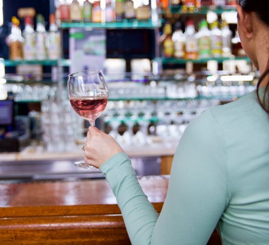 A woman with long dark hair, wearing a light green top, is sitting at a bar holding a glass of red wine. Behind her, shelves are lined with various bottles of alcohol. The atmosphere is cozy and inviting.