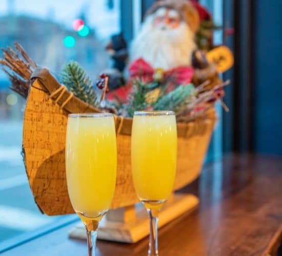 santabrunch Two champagne flutes filled with yellow-orange drinks sit on a wooden windowsill. In the background, there's a decorative Santa figure in a sleigh adorned with pine branches. Street lights and an intersection are visible through the window.